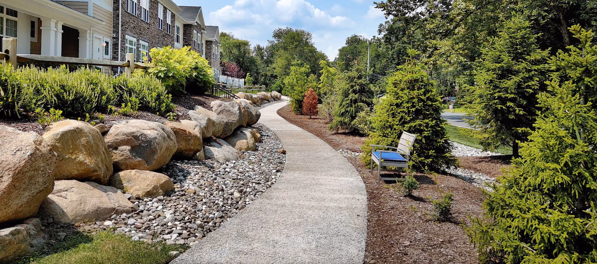 Curved paved walkway bordered by large decorative rocks, landscaped evergreens and a bench beside a multi-unit building on a sunny day.