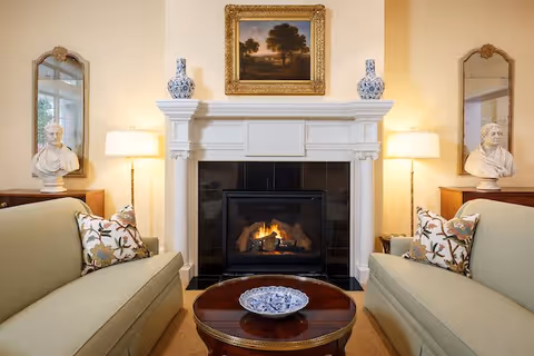 A cozy living room with two beige sofas facing each other, each with floral patterned pillows. Between them is a round wooden coffee table with a decorative blue and white plate. A white fireplace with a fire burning is centered on the back wall, flanked by two table lamps and bust sculptures on wooden side tables. Above the fireplace hangs a framed landscape painting, and two tall mirrors are positioned behind the busts on either side.