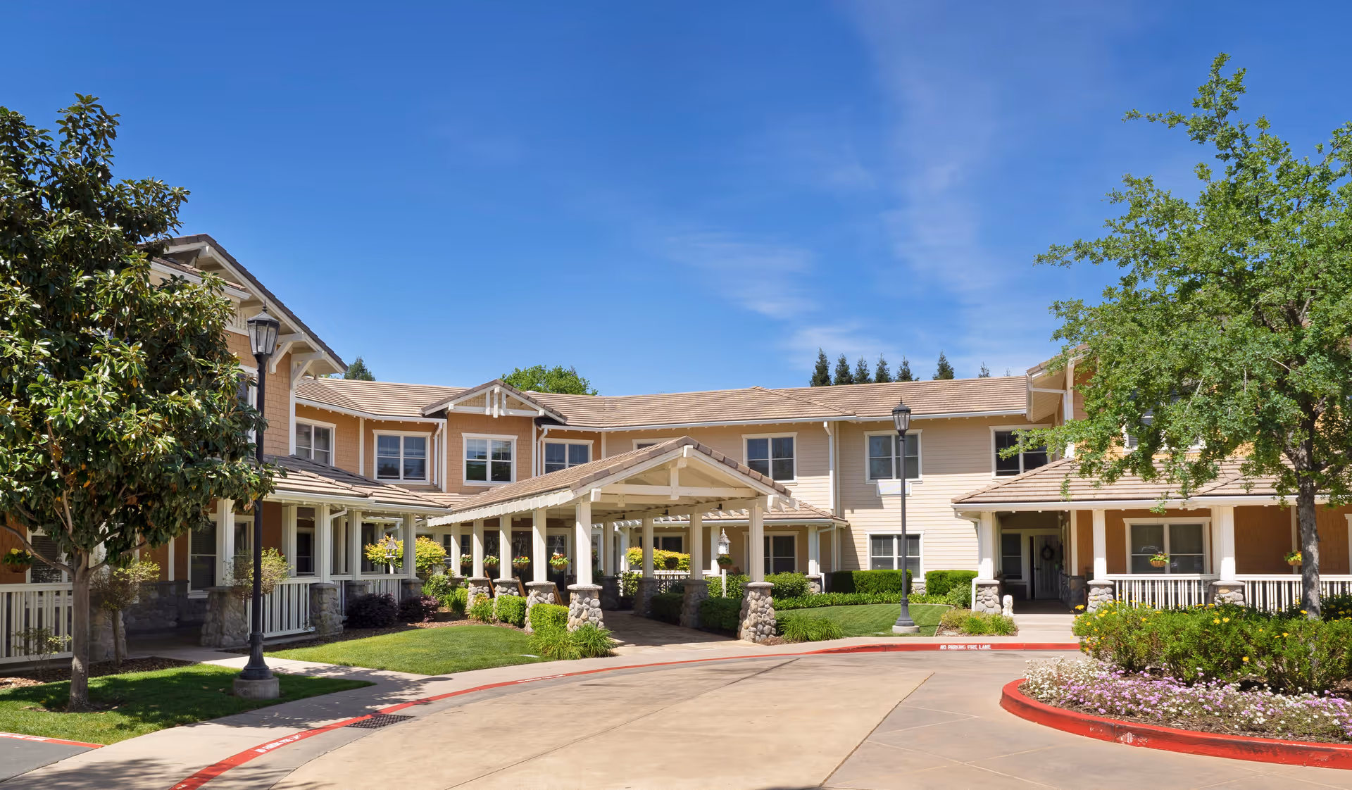 Front exterior of a two-story senior living building with a covered entrance, landscaped grounds, and a clear blue sky.