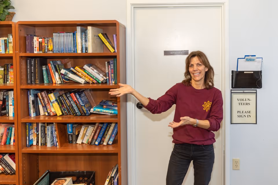 A woman in a maroon long-sleeve shirt and black pants stands smiling and gesturing towards a wooden bookshelf filled with books. Behind her is a white door with a sign that reads 'ACTIVITIES' and a wall-mounted holder with papers and a sign that says 'VOLUNTEERS PLEASE SIGN IN'.
