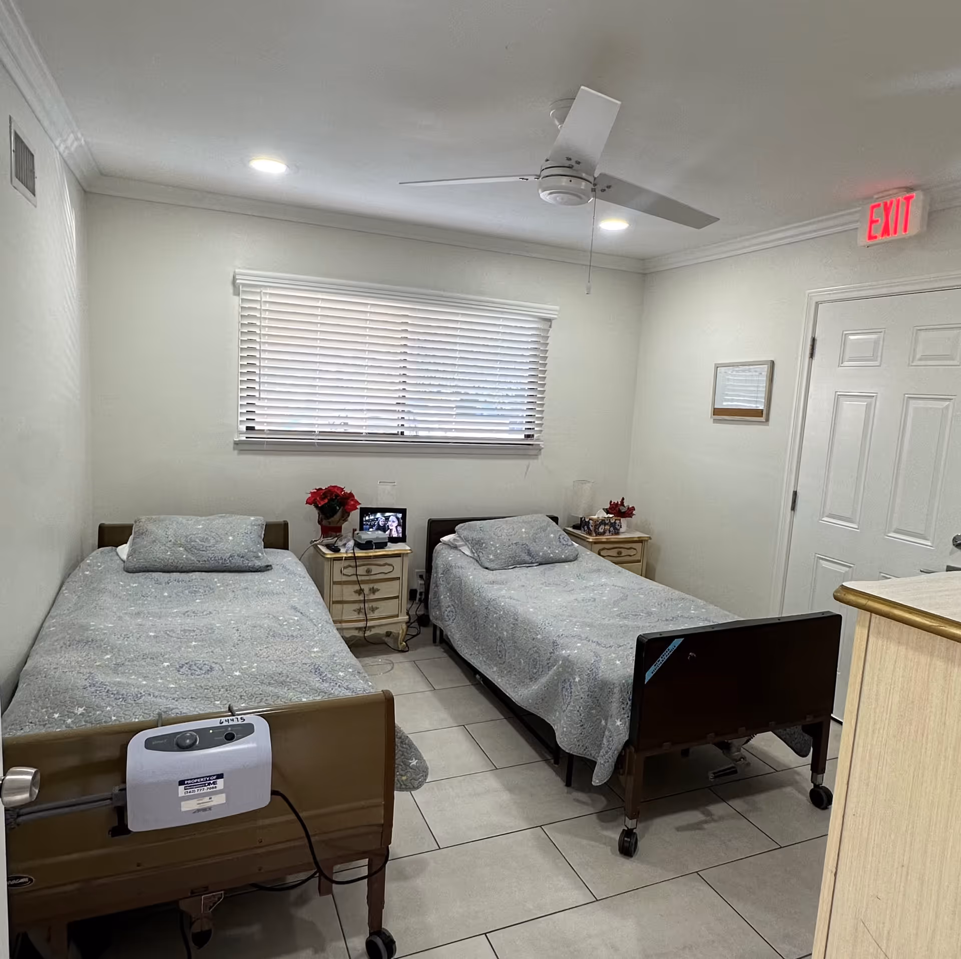 A small bedroom with two single beds, each with a light blue patterned bedspread and a pillow. Between the beds are two nightstands with red flowers, a lamp, and a digital photo frame. The room has white walls, a window with closed blinds, a ceiling fan, tiled floor, and an exit sign above a closed white door.