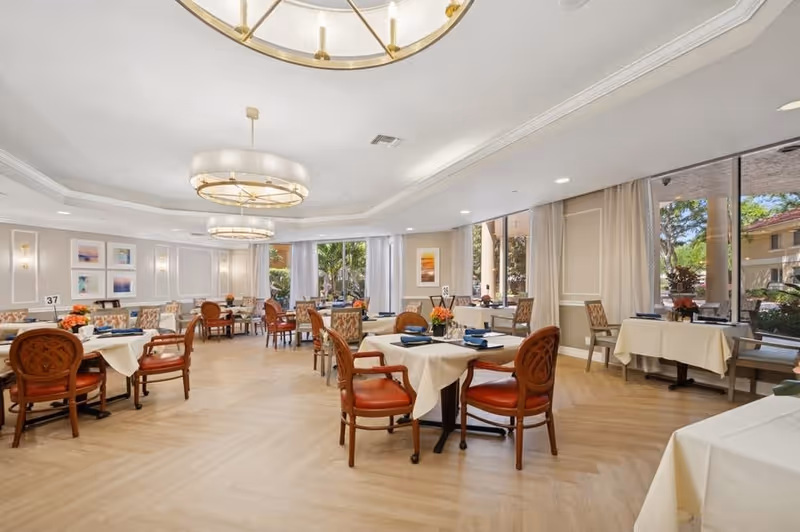 Interior view of a dining room at Five Star Premier Residences of Boca Raton, featuring elegant tables with white tablecloths, wooden chairs, and large windows allowing natural light.