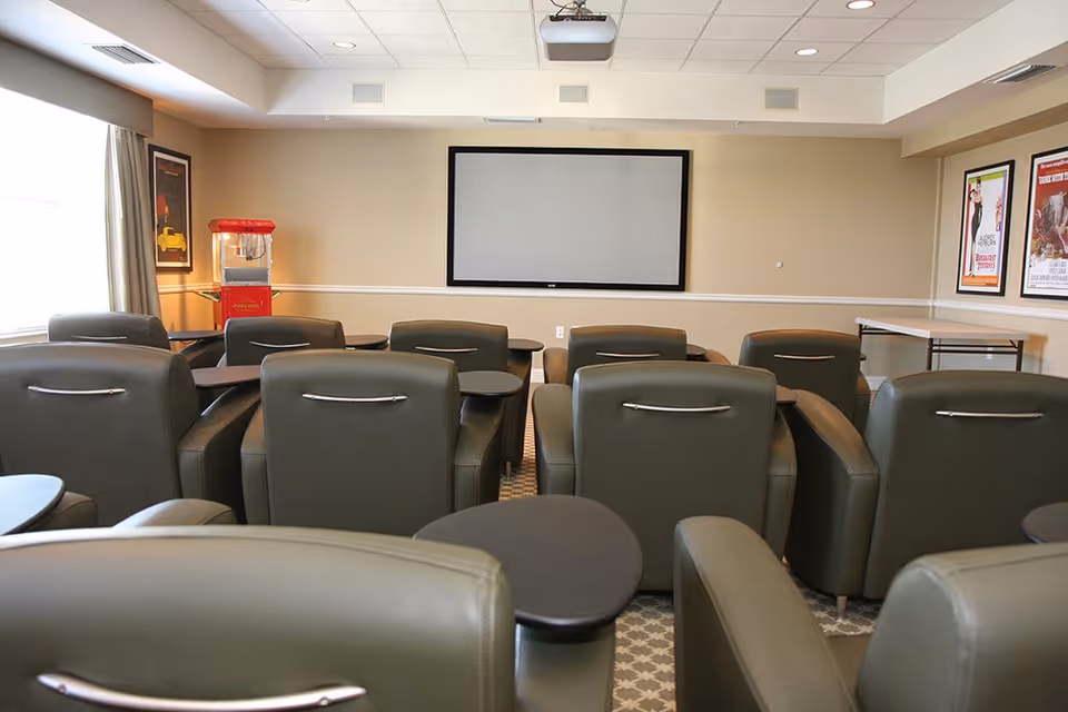 A small theater room with several rows of green leather recliner chairs, each equipped with a small attached table. A large projector screen is mounted on the beige wall at the front. There is a red popcorn machine on a table to the left near a window with curtains, and framed movie posters are hung on the walls.