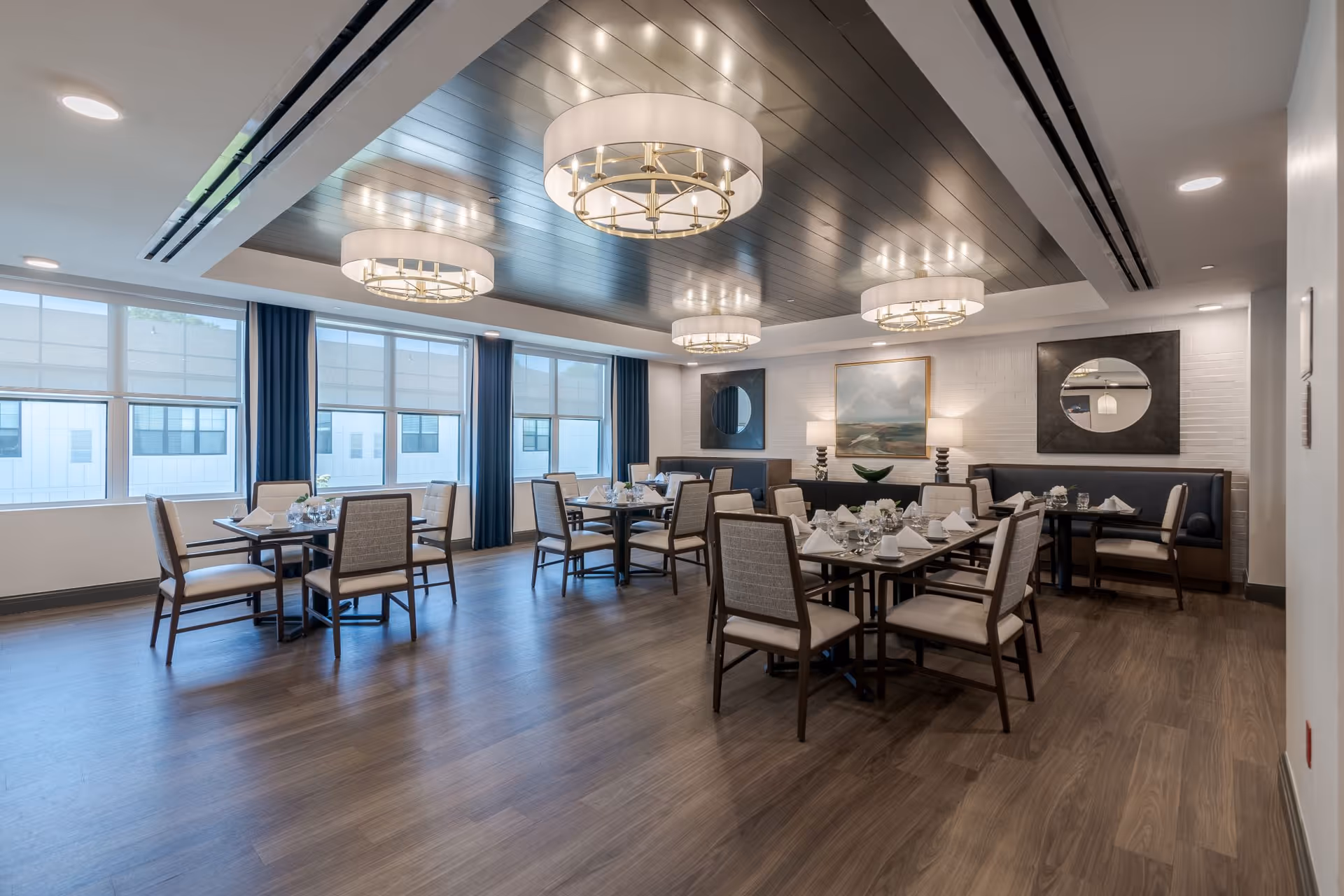 A spacious, well-lit dining room with multiple tables set for meals, featuring chairs with cushioned seats and backs. The room has large windows with blue curtains, modern circular ceiling lights, wood flooring, and decorative wall art including mirrors and a painting.