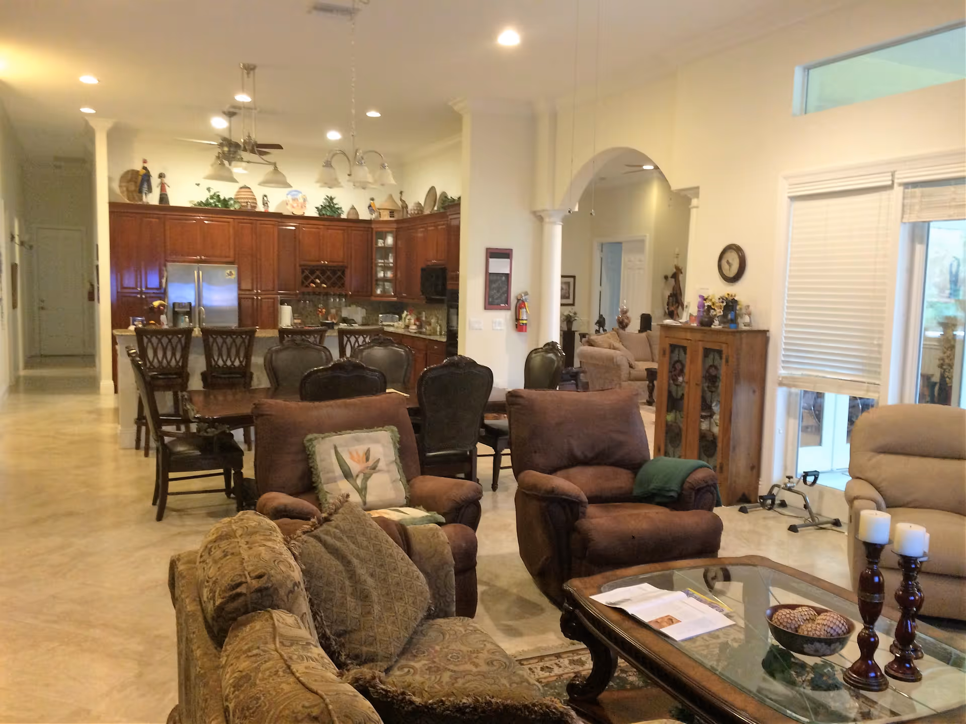 A spacious open-concept living area featuring a seating area with patterned and brown upholstered chairs and sofas, a glass coffee table with decorative items, and a dining area with a wooden table and chairs. The background shows a kitchen with dark wood cabinets, stainless steel refrigerator, and various decorative items on top of the cabinets. The room has beige tiled floors, cream-colored walls, and recessed lighting.