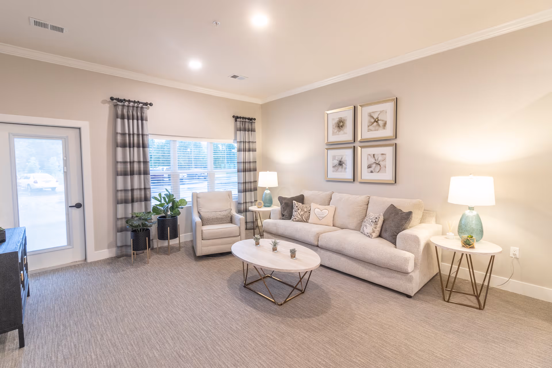 A bright and cozy living room with a beige sofa adorned with multiple cushions, a matching armchair, a round coffee table with small plants, two side tables each with a lamp, and a window with striped curtains letting in natural light.