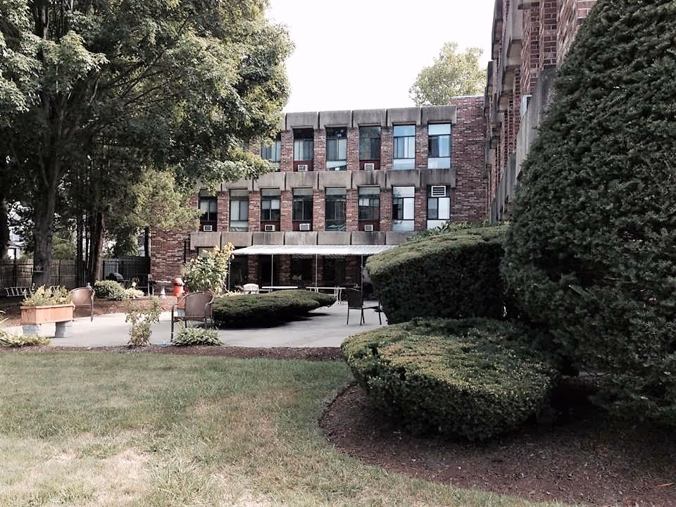 Outdoor courtyard with patio seating, trimmed shrubs, and trees in front of a three-story brick rehabilitation center.