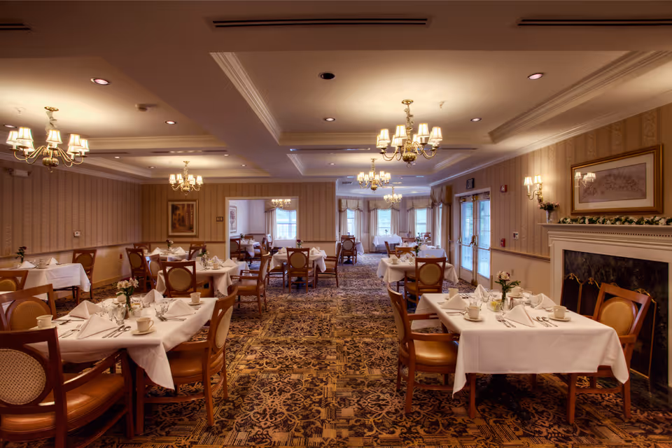 A warmly lit dining room with multiple tables covered in white tablecloths, each set with cups, napkins, and silverware. The room features patterned carpet, wooden chairs with cushioned seats, chandeliers hanging from the ceiling, and a fireplace on the right side with a framed picture above it. Large windows and glass doors allow natural light to enter the space.