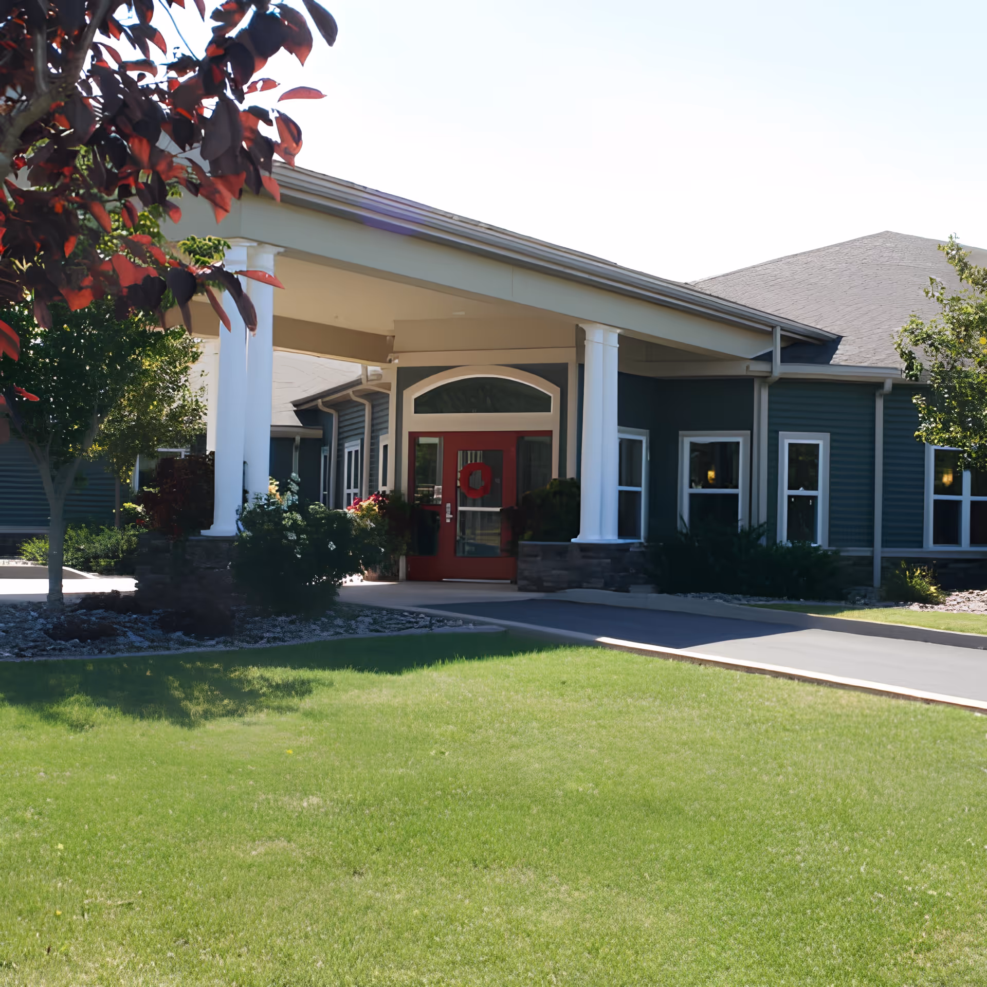 Exterior view of the entrance to a senior living community building with a covered driveway supported by white columns, a red door with a wreath, surrounded by green grass, trees, and shrubs.
