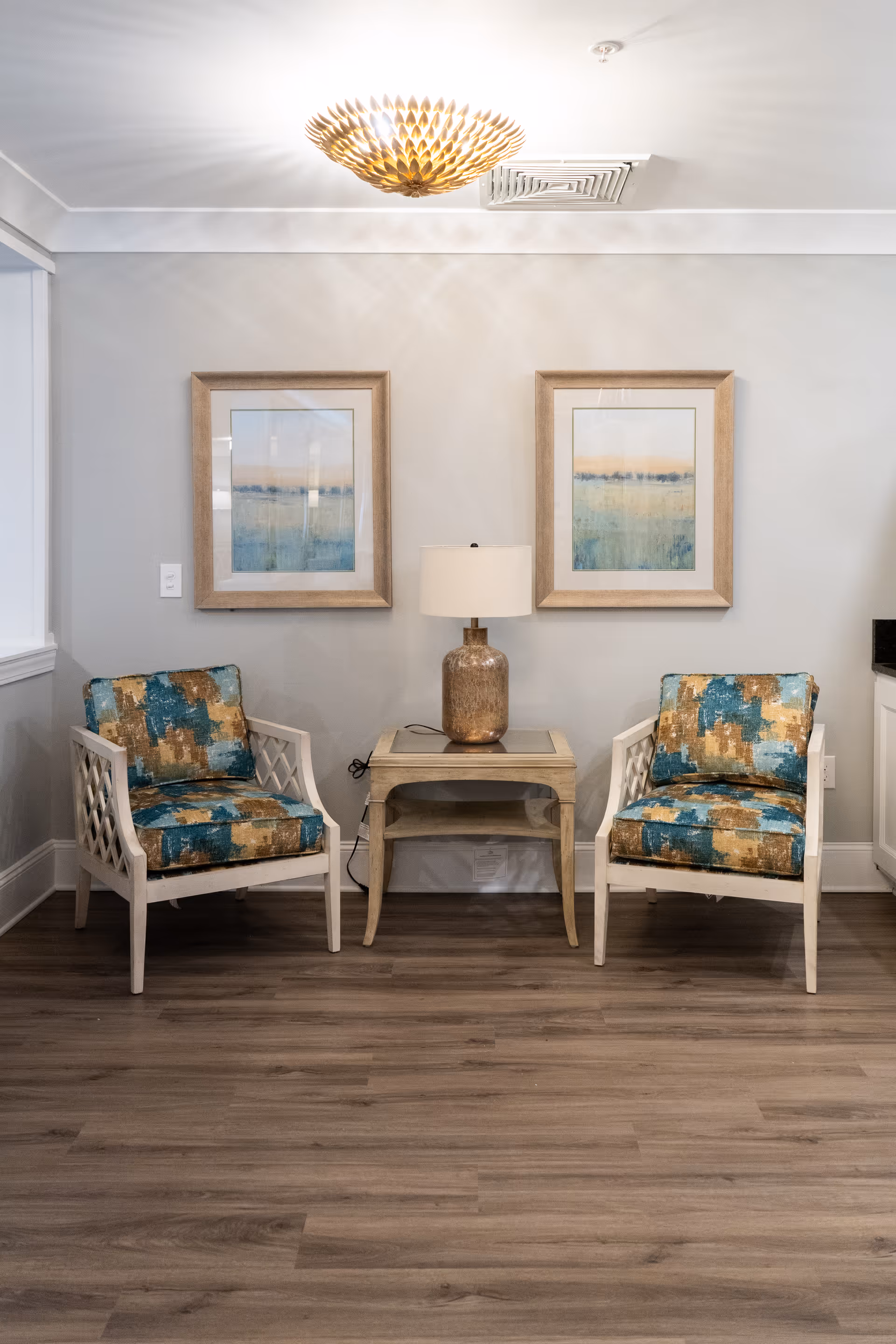 A cozy interior seating area with two cushioned armchairs featuring a blue, gold, and beige pattern. Between the chairs is a small wooden side table with a decorative lamp. Above the table are two framed landscape paintings with soft blue and beige tones. The room has light gray walls, wood flooring, and a decorative ceiling light fixture.