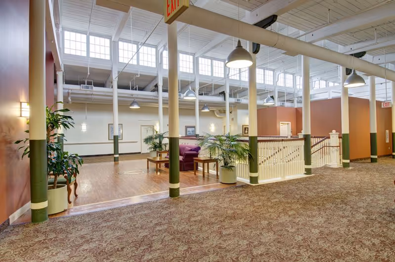 Spacious, well-lit assisted living common area with seating, potted plants, white columns, high windows and pendant lights.