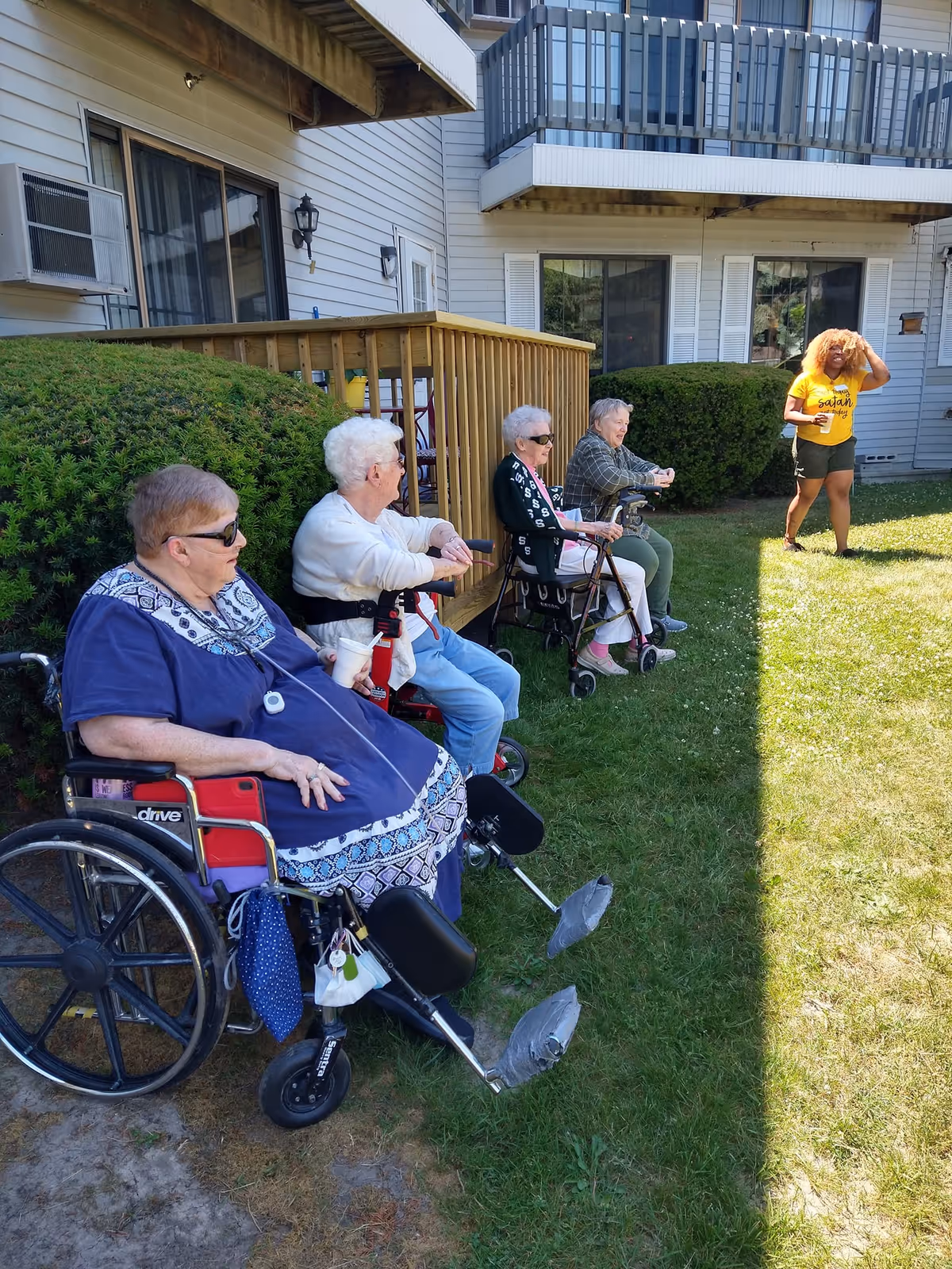 Four elderly women sitting outside on a grassy area near a building, three of them using walkers or wheelchairs, and a younger woman standing in front of them wearing a yellow shirt and shorts.