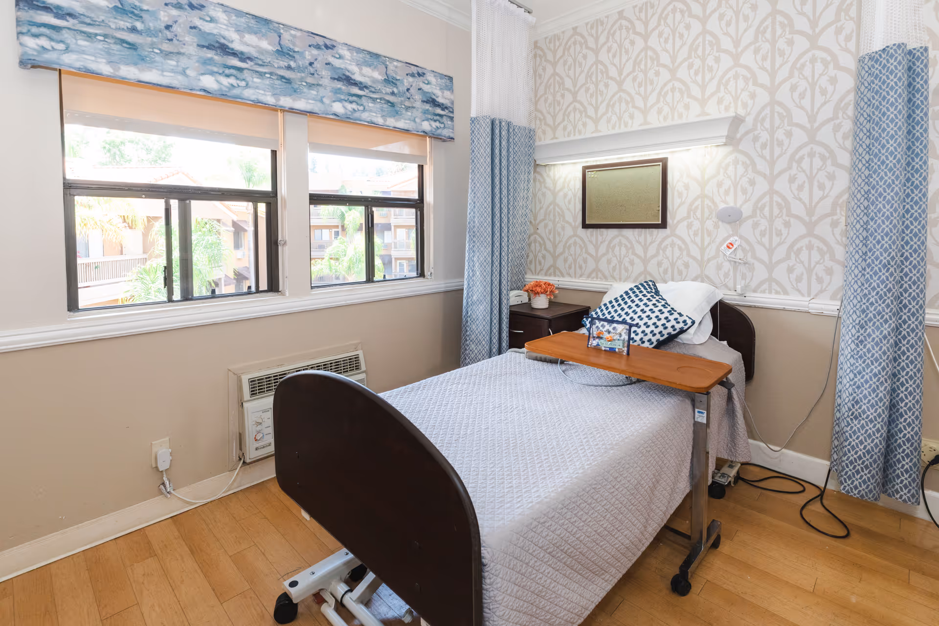 A single bed in a healthcare facility room with a light gray quilted blanket and a blue patterned pillow. There is a wooden overbed table on wheels positioned over the bed. The room has two windows with a blue and white valance and blue patterned curtains. A small nightstand with a potted plant and a framed picture is next to the bed. The walls have a beige and white patterned wallpaper with a white chair rail. The floor is light wood, and there is an air conditioning unit below the windows.