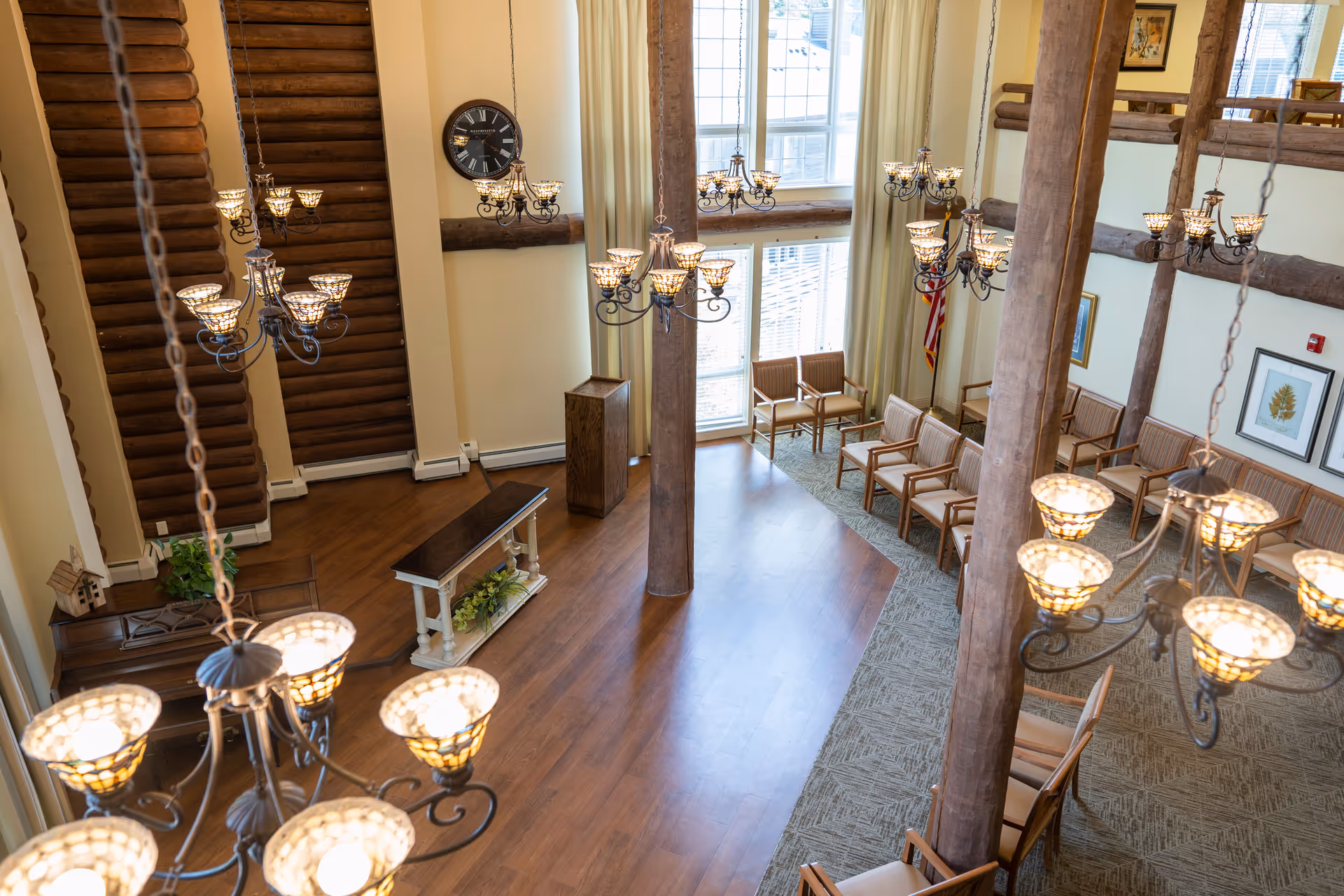 Interior view of a spacious room with high ceilings and large windows allowing natural light. The room features wooden beams and log cabin style walls, multiple elegant chandeliers hanging from the ceiling, rows of wooden chairs arranged facing a small stage area with a podium and a table decorated with plants. There is a large clock on the wall and framed artwork, with an American flag in the corner.