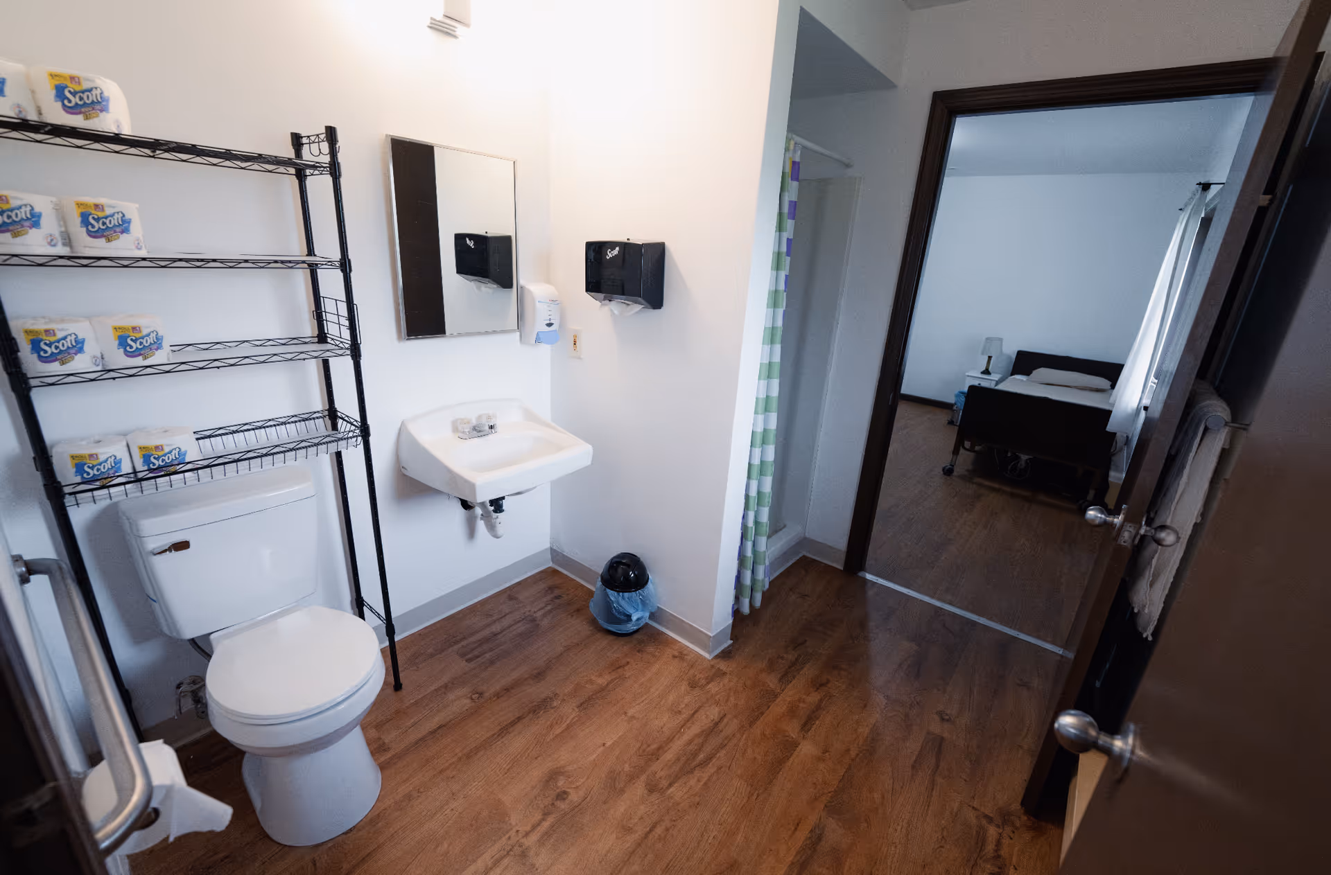 A clean bathroom with a white toilet, a black metal shelving unit stocked with Scott toilet paper, a wall-mounted sink with a mirror above it, a soap dispenser, and a paper towel dispenser. The bathroom has wood-style flooring and white walls. An open door leads to a bedroom with a bed, nightstand, and lamp visible.