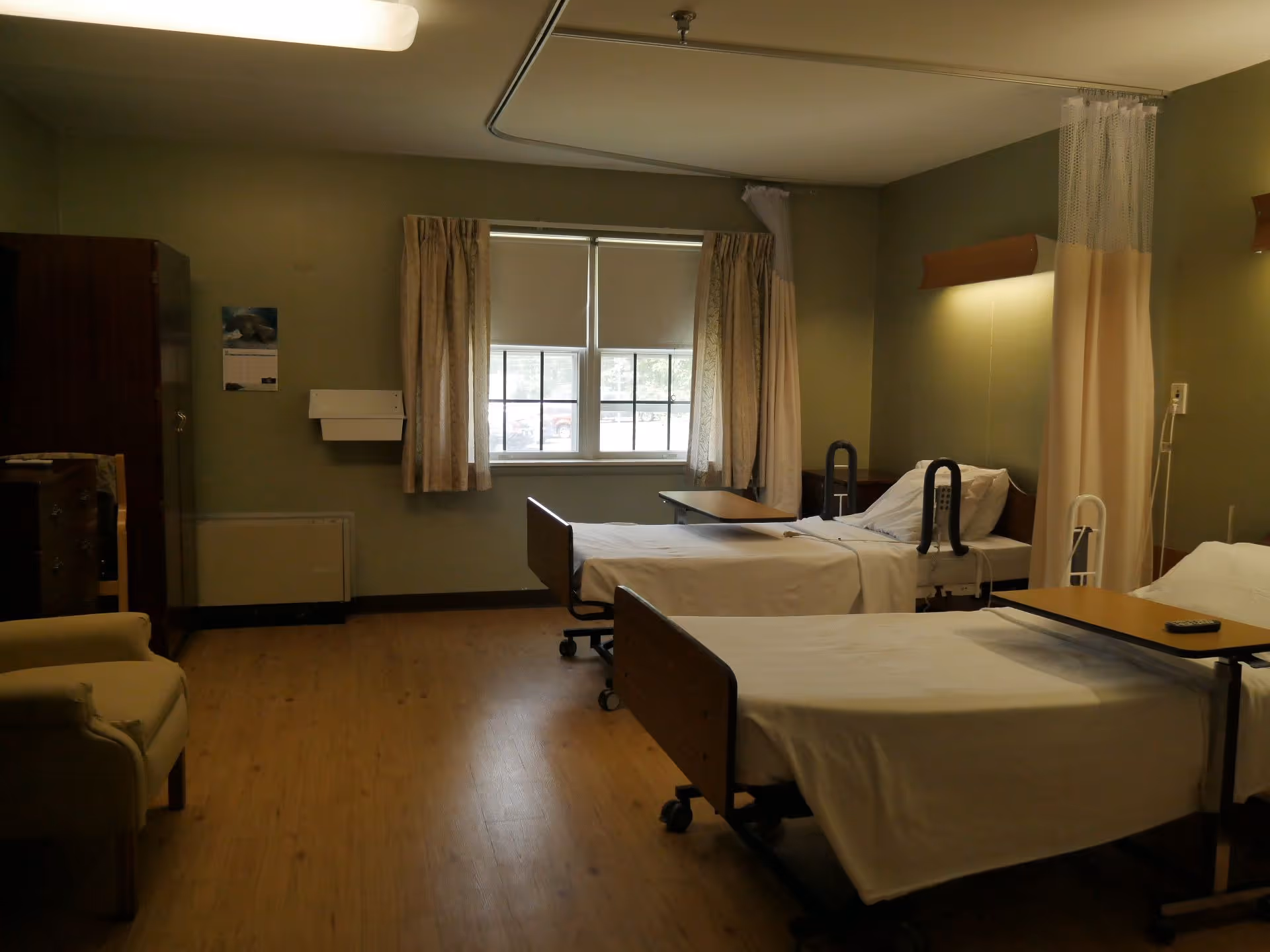 A healthcare facility room with two hospital beds, each equipped with overbed tables and safety rails. The room has green walls, a window with curtains, wooden flooring, a beige armchair, and privacy curtains between the beds.