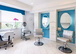 Bright salon room with three styling chairs, oval mirrors set into teal accent walls, and a large window letting in natural light.