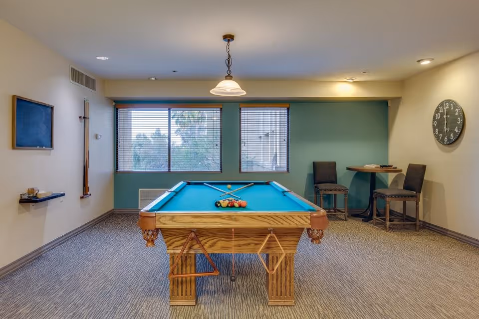 A recreational room with a pool table in the center, pool cues mounted on the wall to the left, a small table with two chairs on the right, a large clock on the wall, and two windows with blinds letting in natural light.
