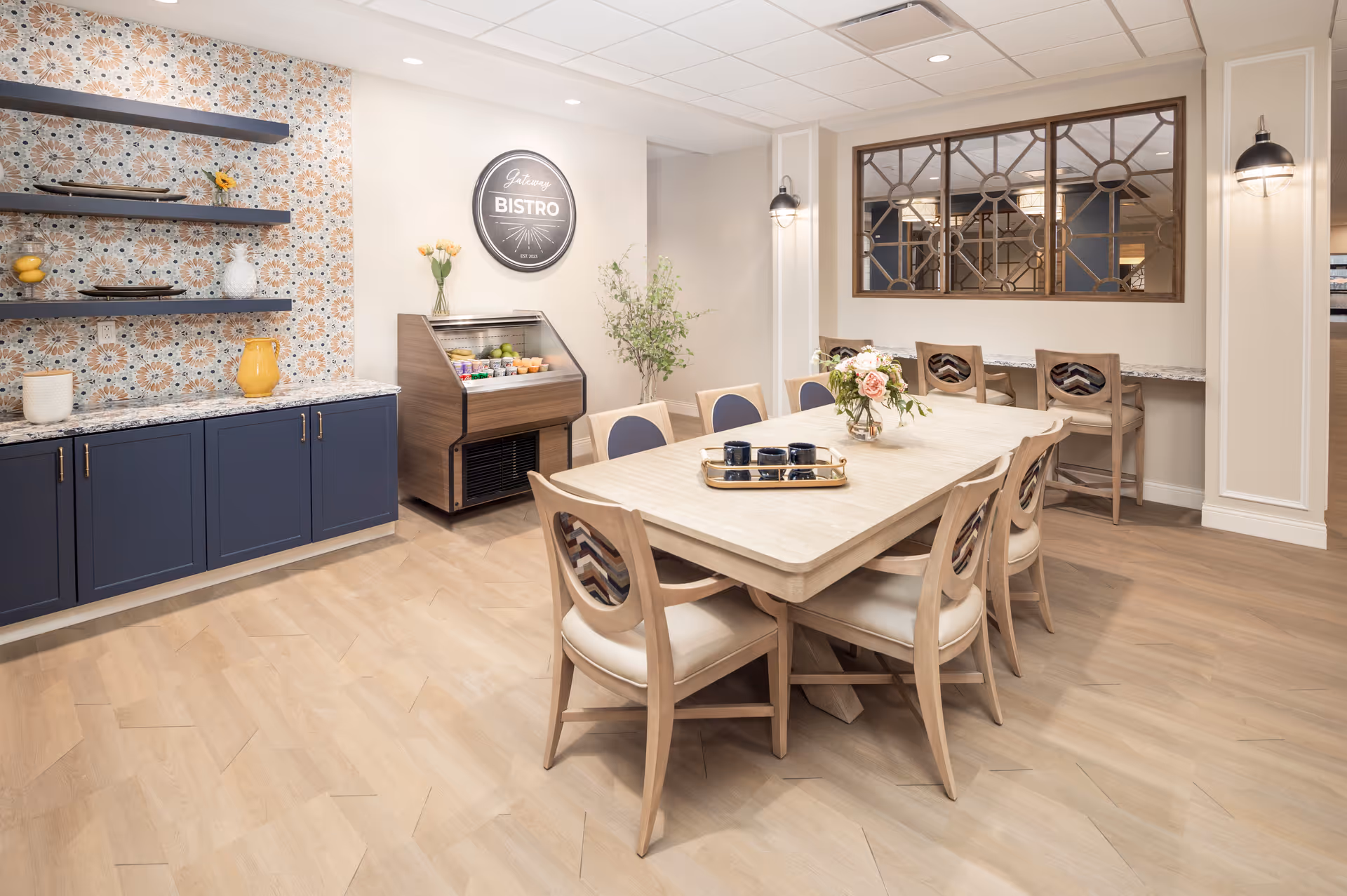 A bright and modern dining area in a senior living facility featuring a large wooden table with eight chairs, a decorative tray with cups, and a floral centerpiece. The room has light wood flooring, a patterned accent wall with shelves holding decorative items, a small refrigerated display case with fruit, and a sign that reads 'Gateway Bistro'. There are also two chairs at a counter beneath a decorative window with geometric metal framing.