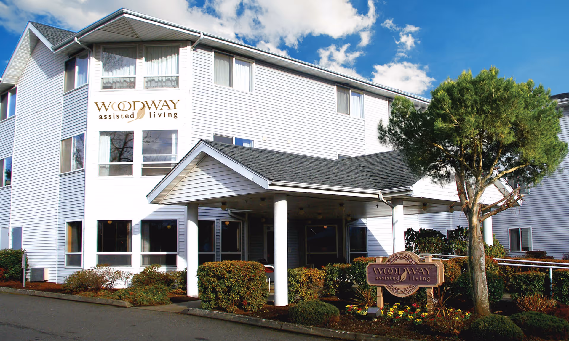 Exterior view of Woodway Senior Living assisted living facility showing a white multi-story building with multiple windows, a covered entrance supported by white columns, landscaped bushes, a tree, and a sign with the facility's name.