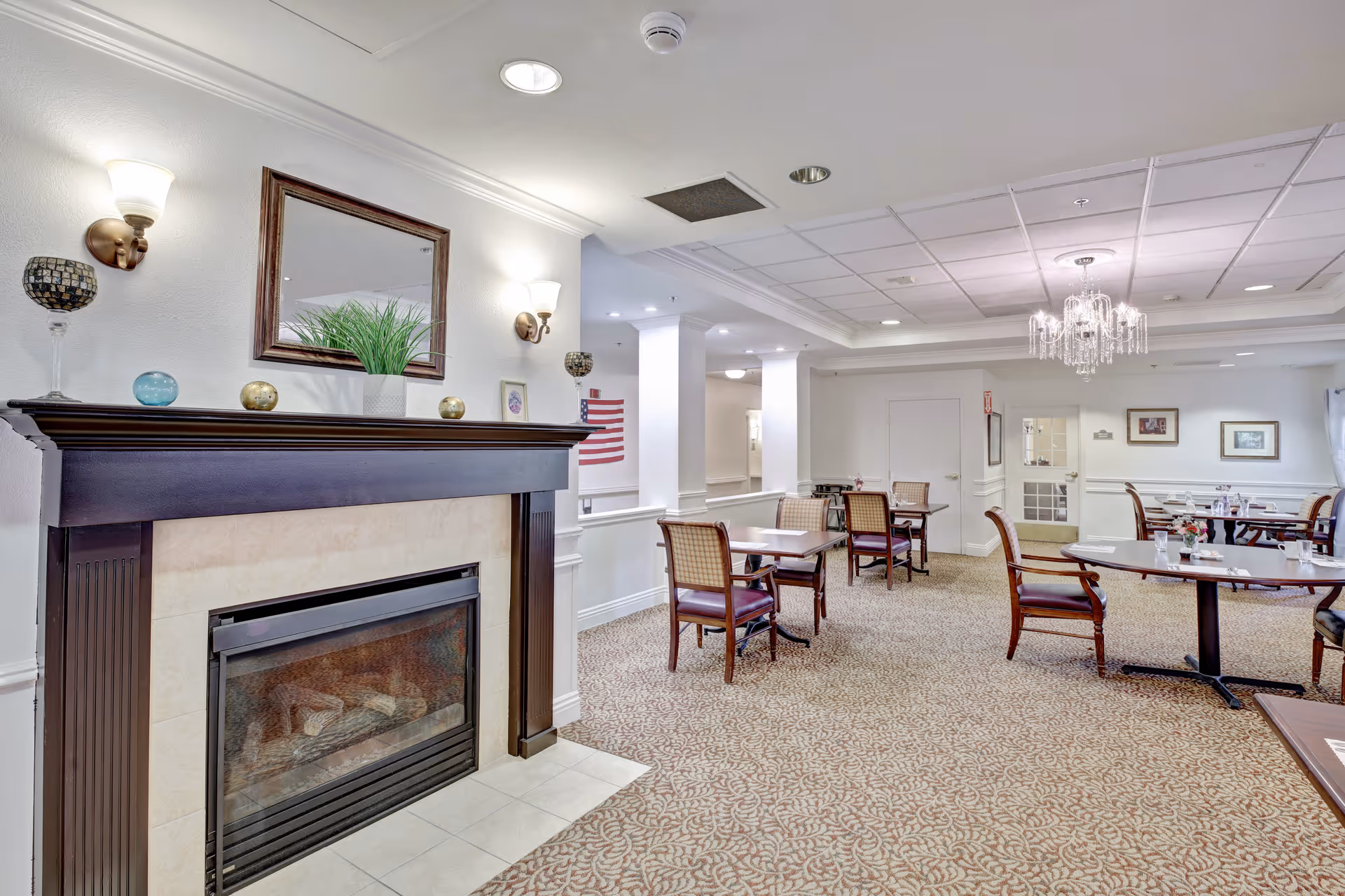 A dining room with several wooden tables and chairs arranged on a patterned carpet. There is a fireplace with a dark wooden mantle decorated with small ornaments and a potted plant. The walls are white with wall sconces providing light, and a chandelier hangs from the ceiling. The room has a calm and inviting atmosphere.