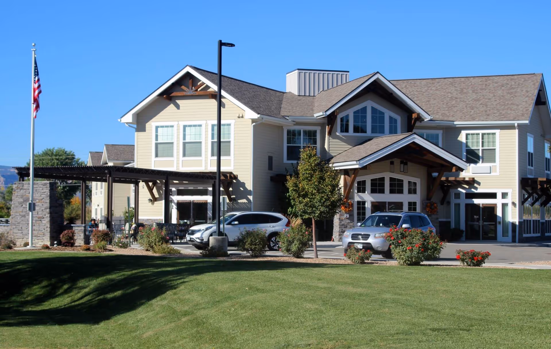 Exterior view of a two-story senior living facility building with beige siding and a gabled roof. There are several windows, a covered entrance with wooden beams, and two cars parked in front. The foreground features a well-maintained lawn, bushes with red flowers, a tree, and an American flag on a flagpole. The sky is clear and blue.