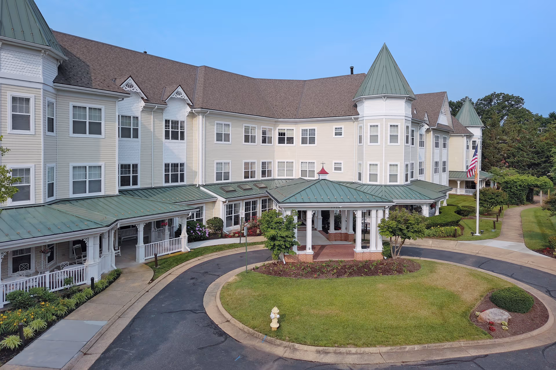 Exterior view of Sunrise of Alexandria, a large senior living facility with beige siding, green metal roofs, multiple windows, and a covered entrance with white columns. The building is surrounded by well-maintained landscaping, a circular driveway, and a flagpole with an American flag.