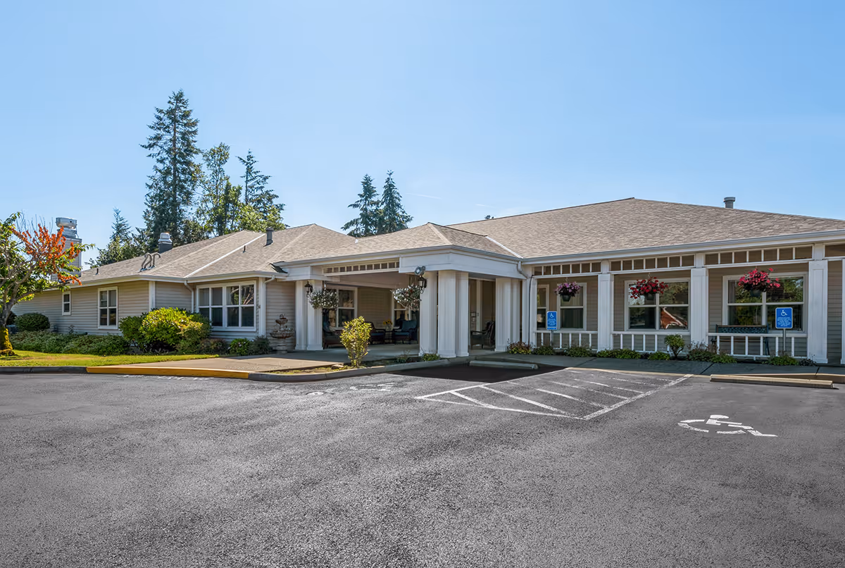 Exterior view of a single-story senior living facility building with a covered entrance, surrounded by greenery and trees under a clear blue sky. There are handicap parking spaces in front of the building.