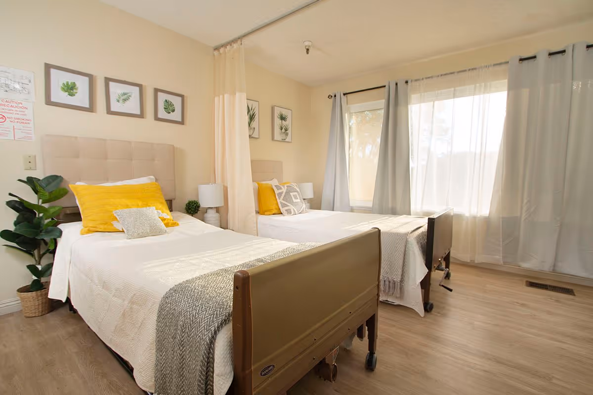 A bright and clean bedroom with two single beds, each with beige headboards, white bedding, yellow and gray pillows, and gray throws. The room has light wood flooring, large windows with sheer and gray curtains, framed botanical prints on the walls, a potted plant, and a privacy curtain between the beds.