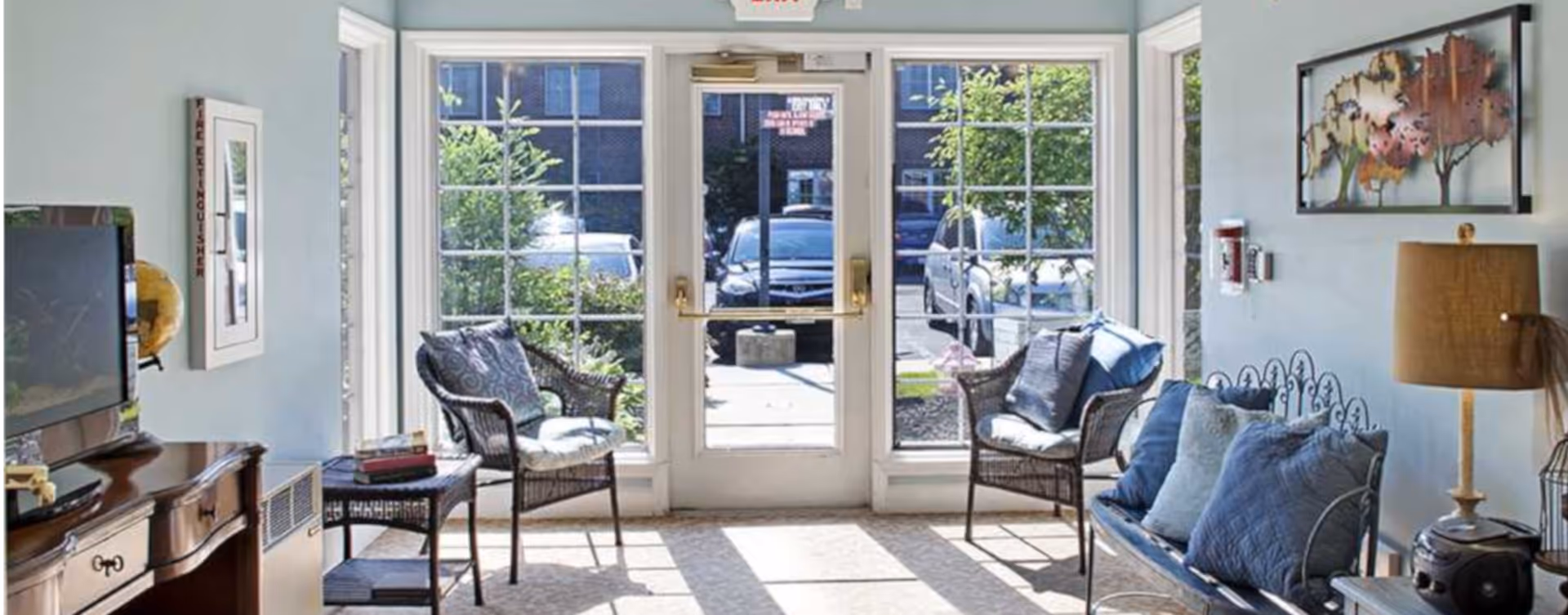 Sunlit seating area/lobby with wicker chairs, a cushioned bench, a TV cabinet, and a glass door with large windows showing parked cars outside.