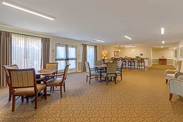 A spacious common area in a senior living facility with multiple seating arrangements including tables with chairs and armchairs. The room has large windows with sheer curtains allowing natural light to enter, beige patterned carpet, and neutral-colored walls. There is a counter with bar stools in the background and framed artwork on the walls.