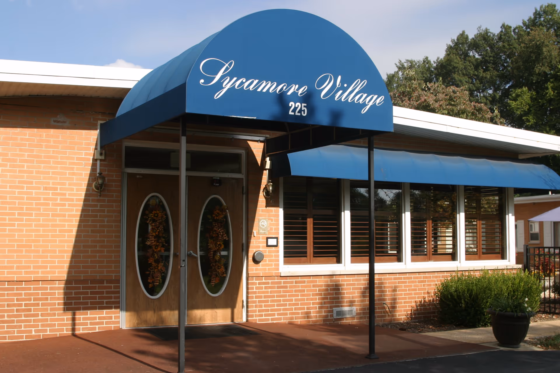 Entrance of Sycamore Village Assisted Living facility with a blue awning displaying the name and number 225 above double wooden doors decorated with oval glass panels and wreaths. The building is made of red brick with several windows covered by a blue awning and surrounded by greenery.