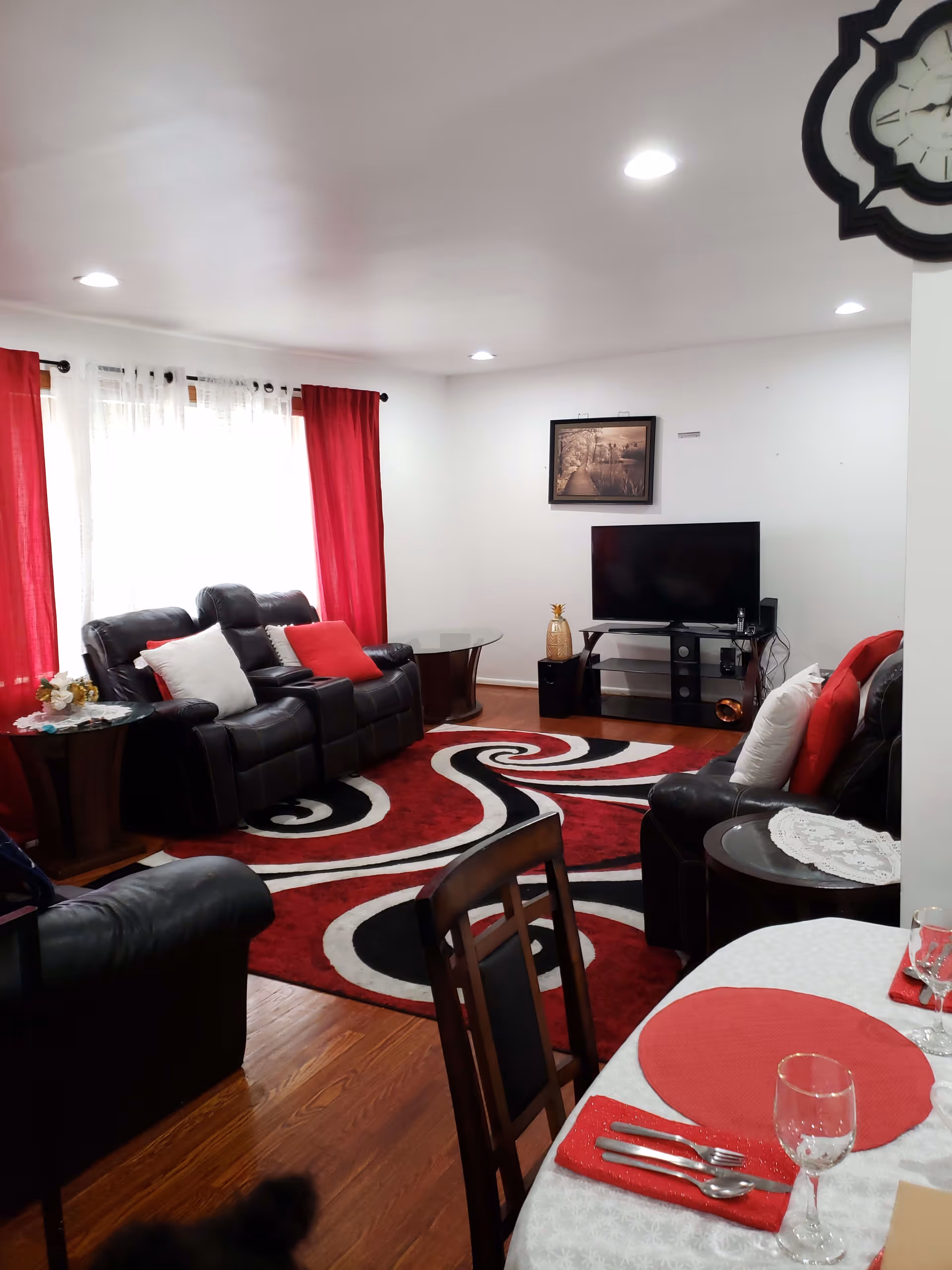 A cozy living room with black leather recliner sofas adorned with red and white pillows, a red, black, and white patterned rug, a flat-screen TV on a black stand, and red curtains on the window. A dining table with a white tablecloth, red placemats, and glassware is partially visible in the foreground. A decorative wall clock and framed artwork are also visible on the walls.
