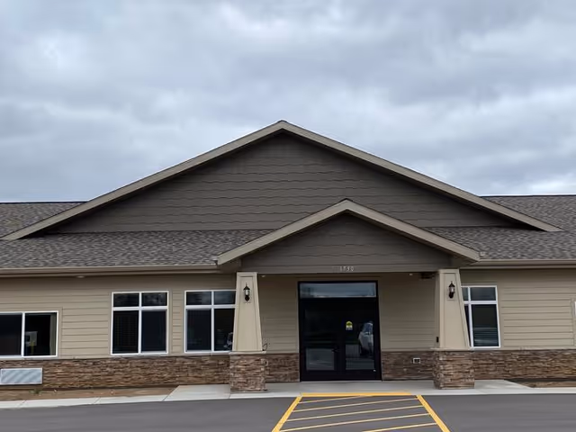 Front exterior view of a single-story building with beige siding and stone accents. The building has a peaked roof with two outdoor wall lanterns flanking a glass entrance door. The sky is overcast and there is a marked parking area in front.