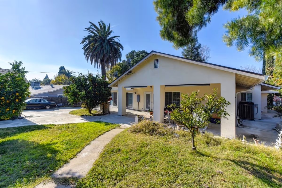 Exterior view of a single-story house with a covered patio area, surrounded by green grass, small trees, and a palm tree in the background under a clear blue sky.