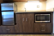 A compact kitchen area featuring a stainless steel refrigerator on the left, wooden cabinets with silver handles below a countertop, and a microwave oven built into the cabinetry on the right. The space is well-lit with a light fixture above the countertop.