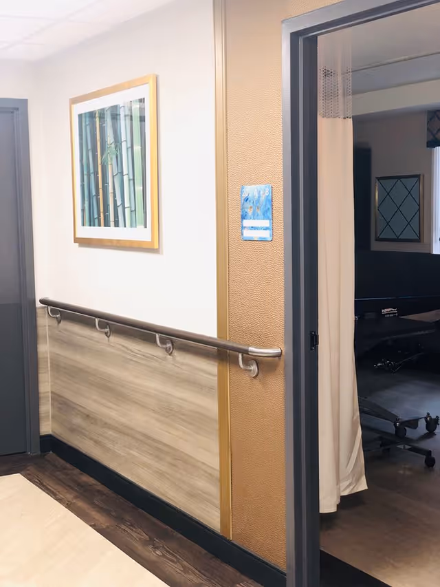 A hallway in a nursing and rehabilitation facility with a handrail along the wall, a framed picture of bamboo on the wall, and an open doorway with a curtain partially drawn revealing a room with a medical bed and a window with a diamond-patterned frame.