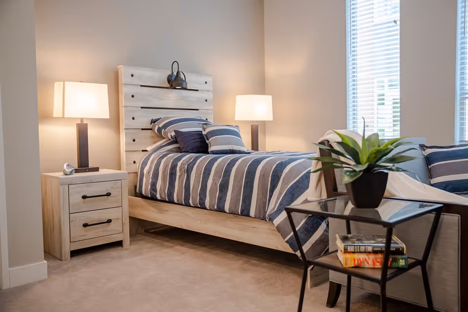 A cozy bedroom with a wooden bed frame and headboard, a striped blue and white bedspread with matching pillows, two bedside lamps on either side, a wooden nightstand with two drawers, and a glass side table holding a potted plant and some books near a window with blinds.