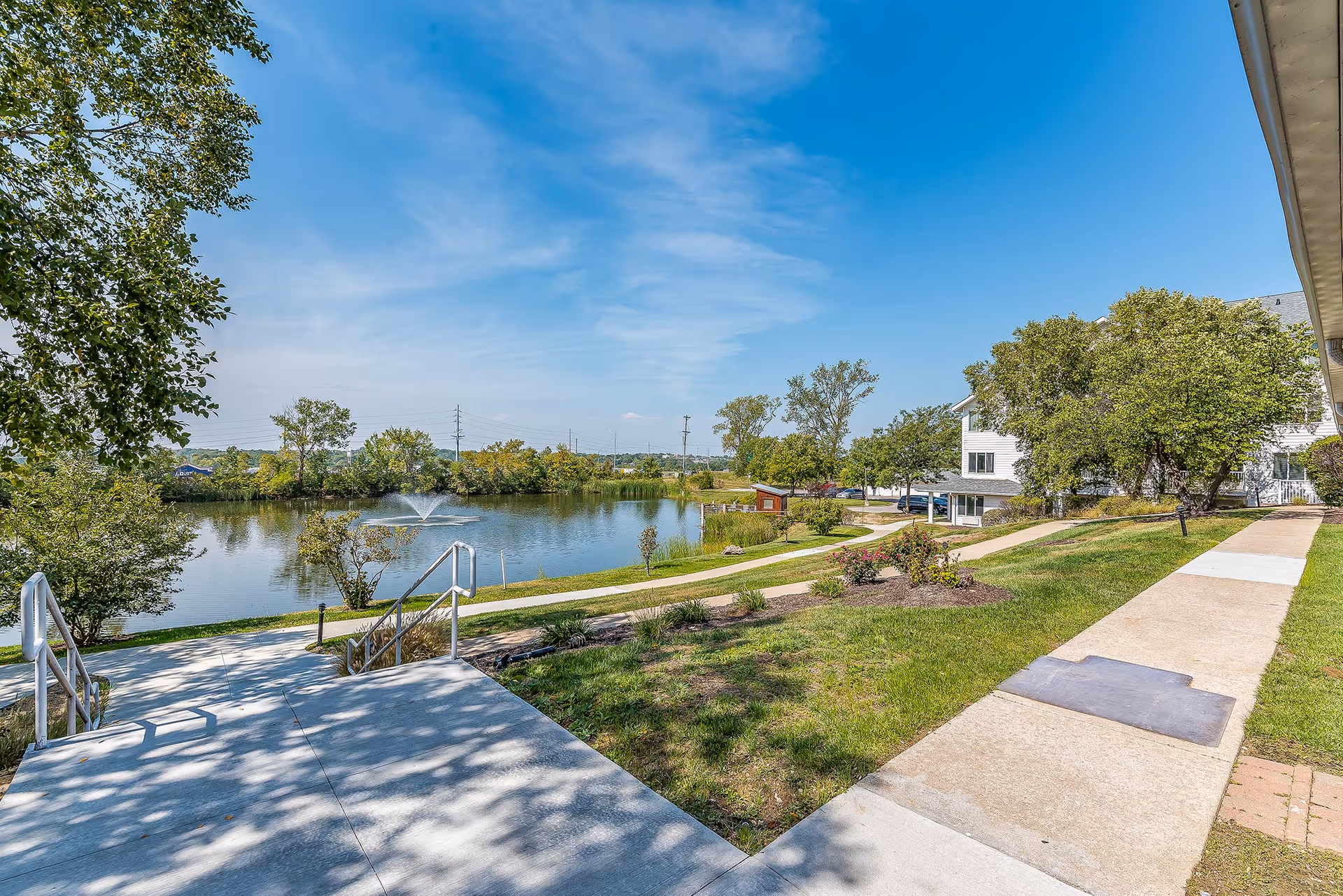 Outdoor view of a senior living facility with a paved walkway, green grass, trees, and a pond with a water fountain under a clear blue sky.
