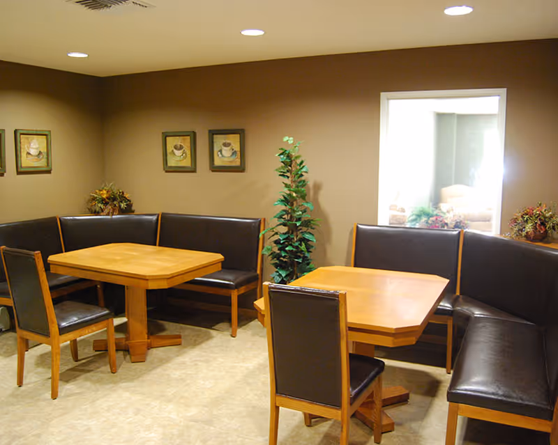 A cozy dining area with two wooden tables surrounded by black cushioned bench seating and chairs. The walls are painted beige and decorated with framed pictures of coffee cups. There are two small plants placed in the corners, and a window on the right side shows a glimpse of another room.