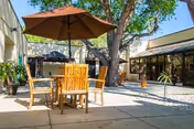 Outdoor patio area with wooden table and chairs under a large brown umbrella, surrounded by trees and buildings with large windows, under a clear blue sky.