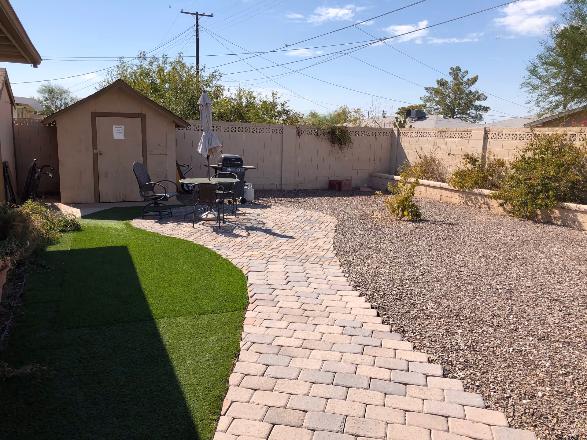 Outdoor patio area with a paved walkway leading to a small round table with three chairs and a closed umbrella. There is a barbecue grill next to the table. A small shed is visible in the background along with a block wall fence and some sparse vegetation and trees. The sky is clear with a few clouds.