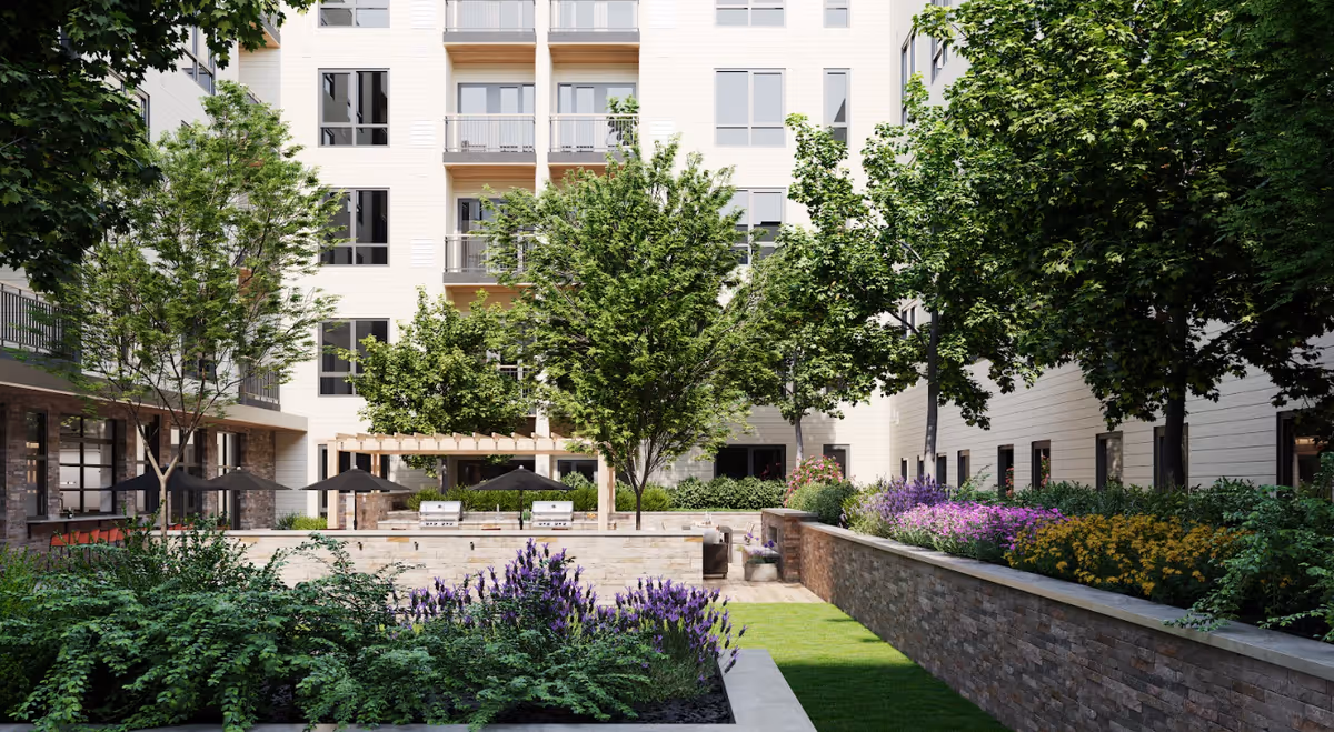 Outdoor courtyard area of a senior living facility with lush green trees, colorful flower beds, stone walls, and a pergola with seating and umbrellas. The multi-story building with balconies surrounds the courtyard.