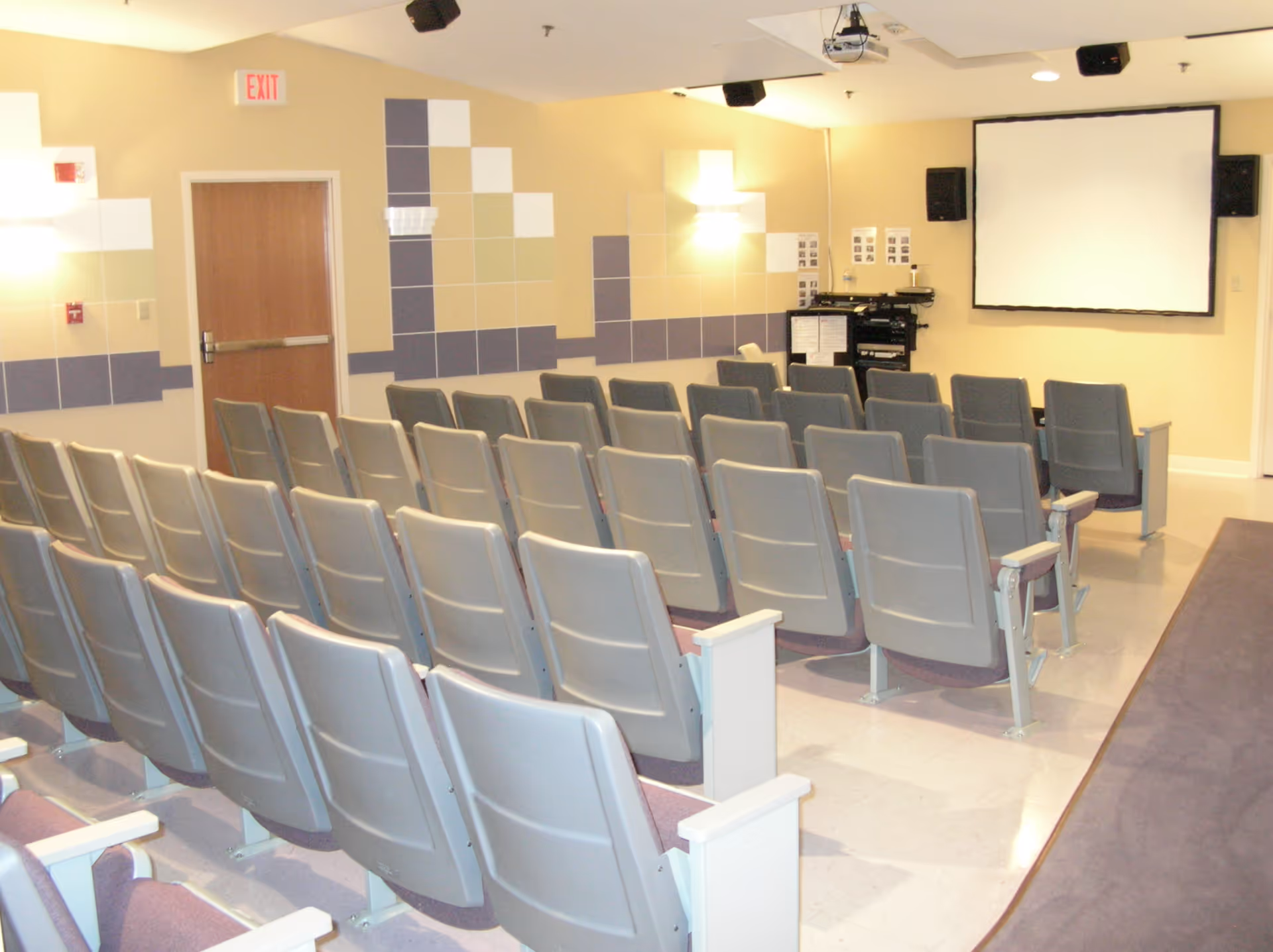 A small auditorium or presentation room with several rows of gray chairs facing a blank projection screen on a beige wall. The room has patterned wall tiles in shades of beige, gray, and white, ceiling-mounted speakers, and a door with an exit sign above it.