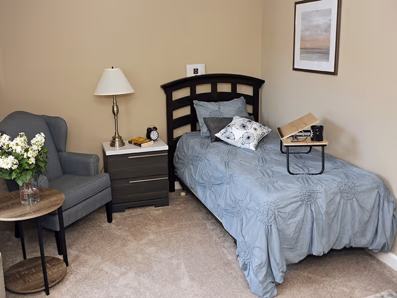 A cozy bedroom with a single bed covered in a blue textured bedspread and several pillows. Next to the bed is a dark wooden nightstand with a lamp, an alarm clock, and a book. A small wooden side table with a vase of white flowers and a gray upholstered armchair are also visible. A framed picture hangs on the beige wall above the bed.
