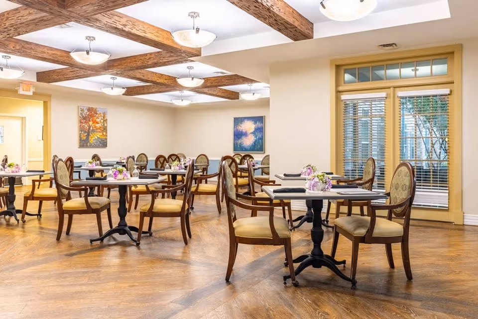 A bright dining room with wooden floors and ceiling beams, featuring several round tables each surrounded by four upholstered chairs. The tables are set with small floral centerpieces and black napkins. The room has large windows with blinds and two paintings on the walls.