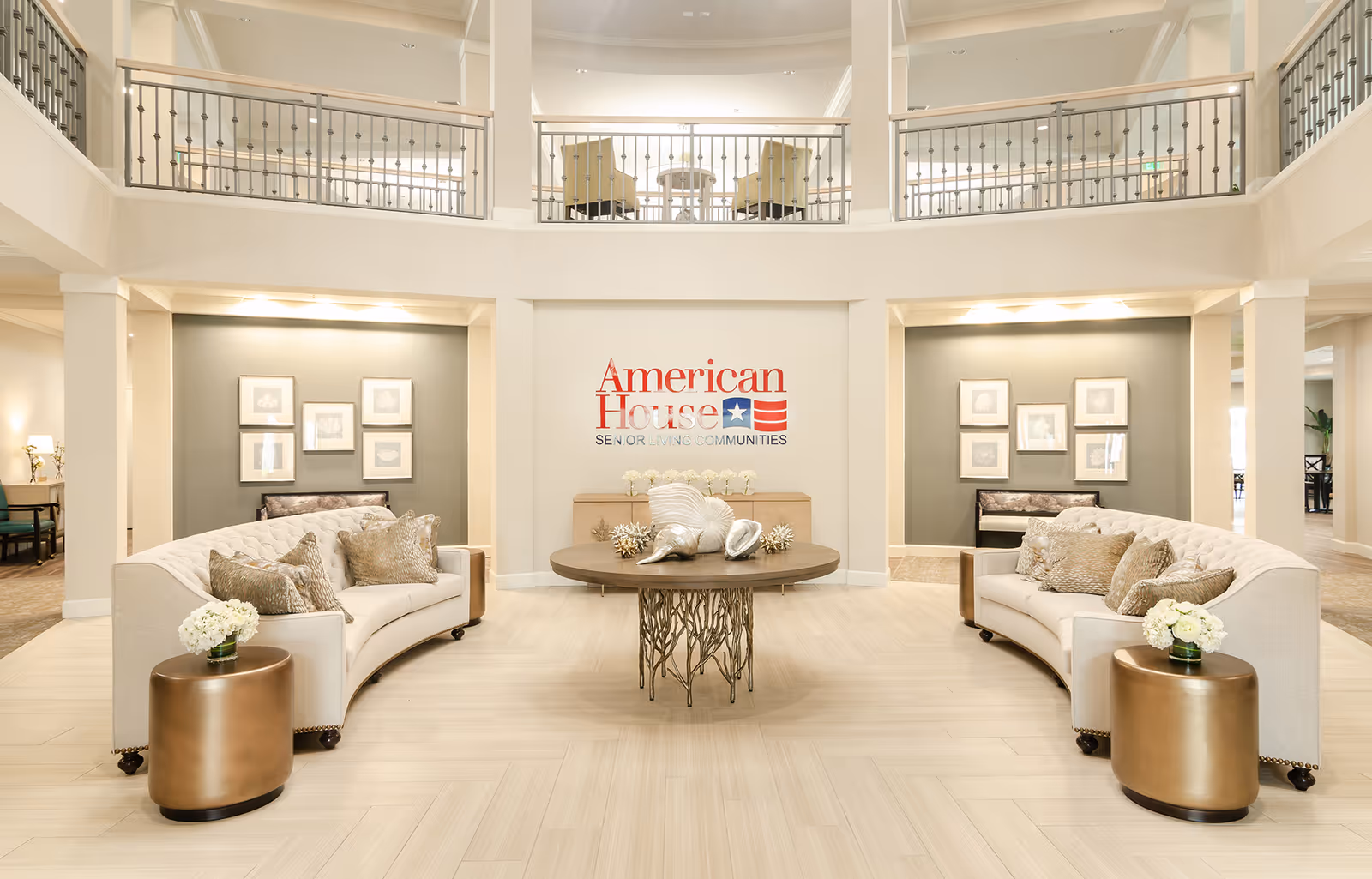 Spacious and elegant senior living community lobby with two curved beige sofas facing each other, a round table with decorative seashells in the center, and a wall sign that reads 'American House Senior Living Communities'. The area is well-lit with soft lighting and features a second-floor balcony with railings overlooking the lobby.