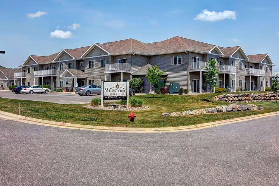 Two-story beige senior living building with balconies, a parking lot and landscaped lawn centered around a Milestone Senior Living sign.