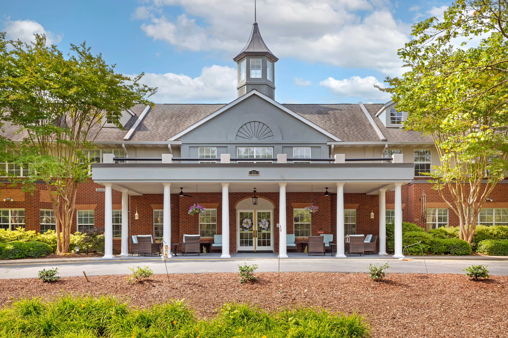 Front exterior view of a senior living facility named Brookdale MacArthur Park, featuring a brick building with white columns supporting a covered entrance. There are outdoor seating areas with chairs and tables under the covered porch, surrounded by green trees and landscaping under a partly cloudy sky.