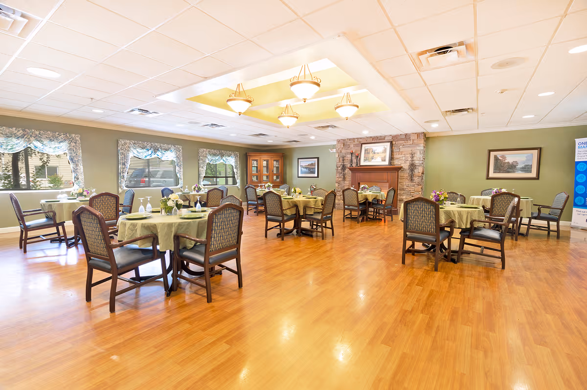 Bright dining room with round tables set with green tablecloths, wooden chairs, hardwood floor and a stone fireplace.